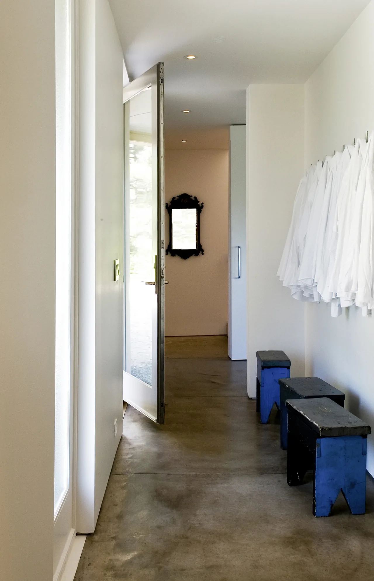 Corridor featuring concrete flooring, white hanging coats, and blue wooden stools along one wall.