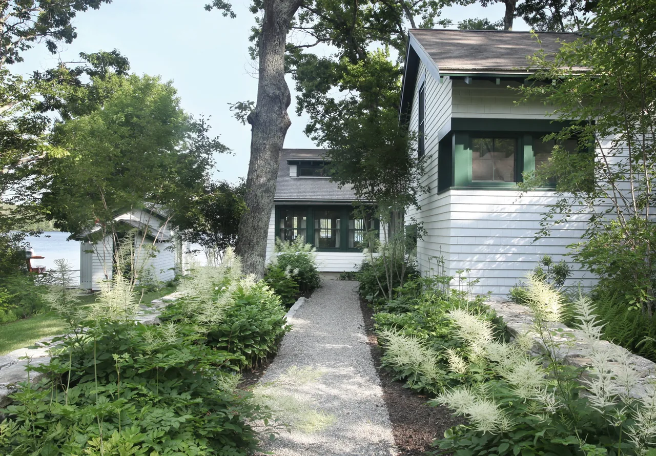 White cottage with dark green window frames sits at the end of a garden path by the lake.