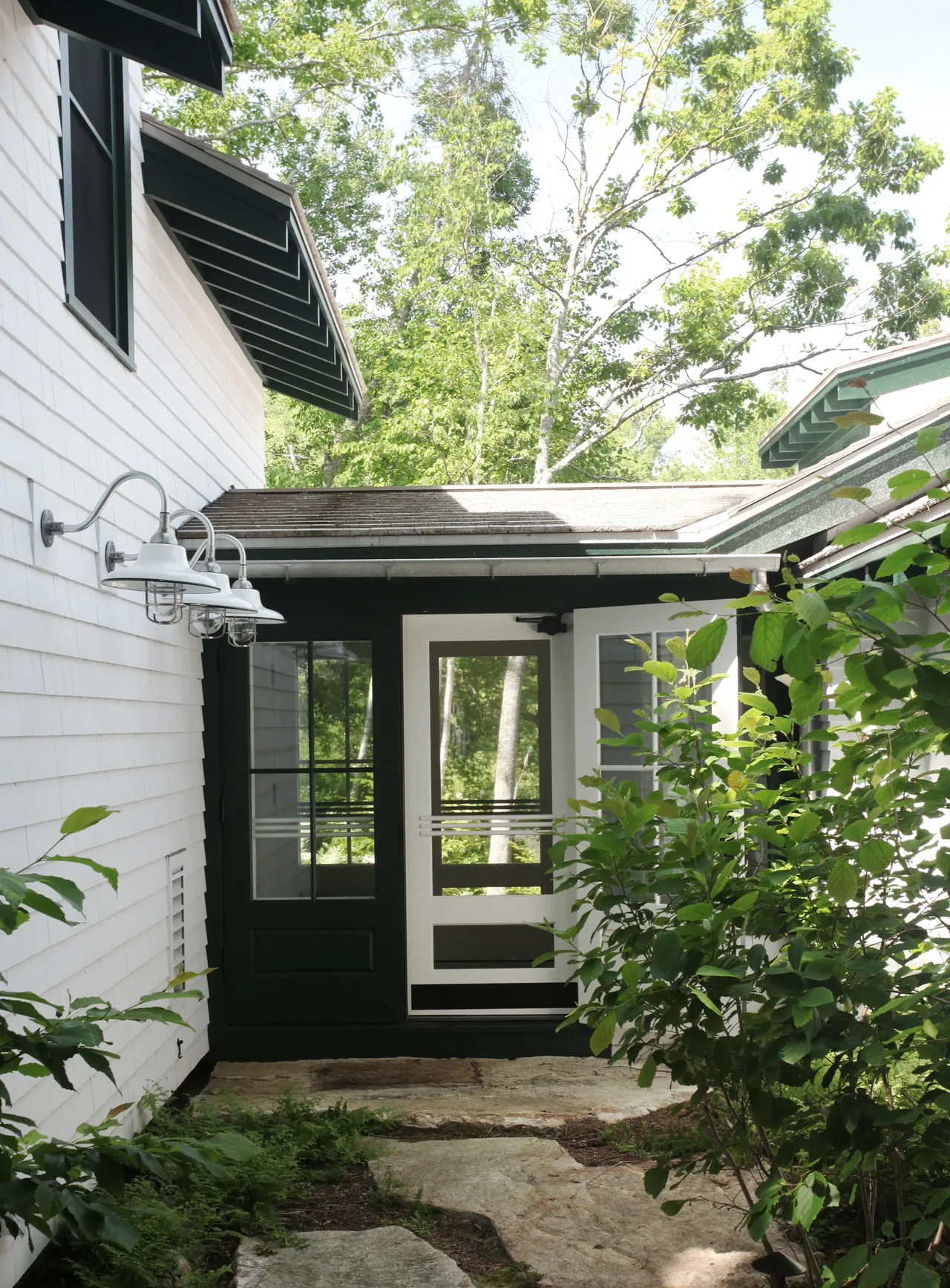 Entryway between two sections of a white shingle-sided house features contrasting black and white glass paneled doors and outdoor wall lights.