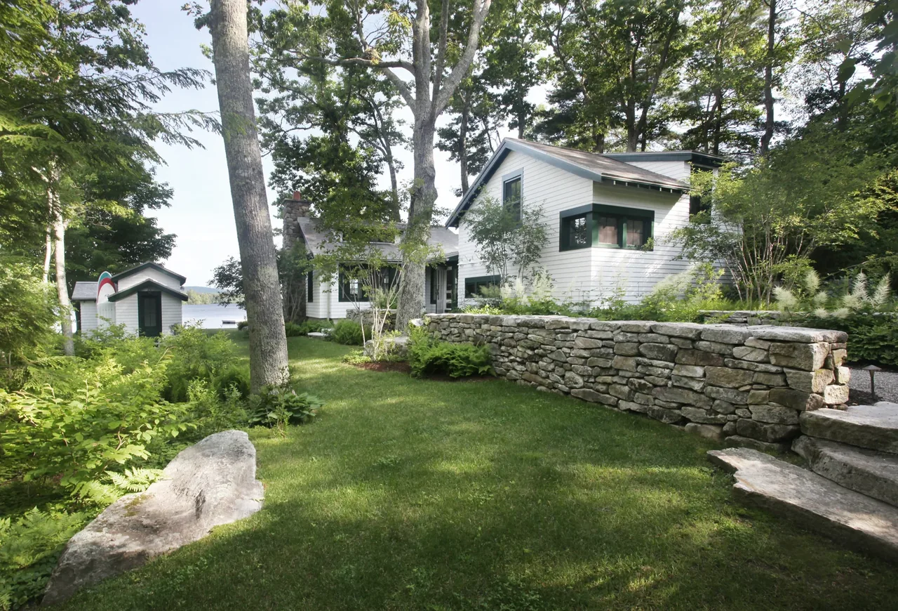 Lakefront cottage with white siding and green window trim, set behind a stone retaining wall and surrounded by trees and lush greenery.
