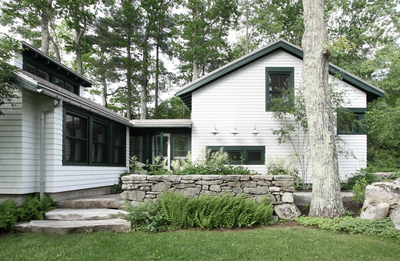 Lakefront cottage with white shingle siding, green window trim, stone retaining walls, and lush garden foliage.