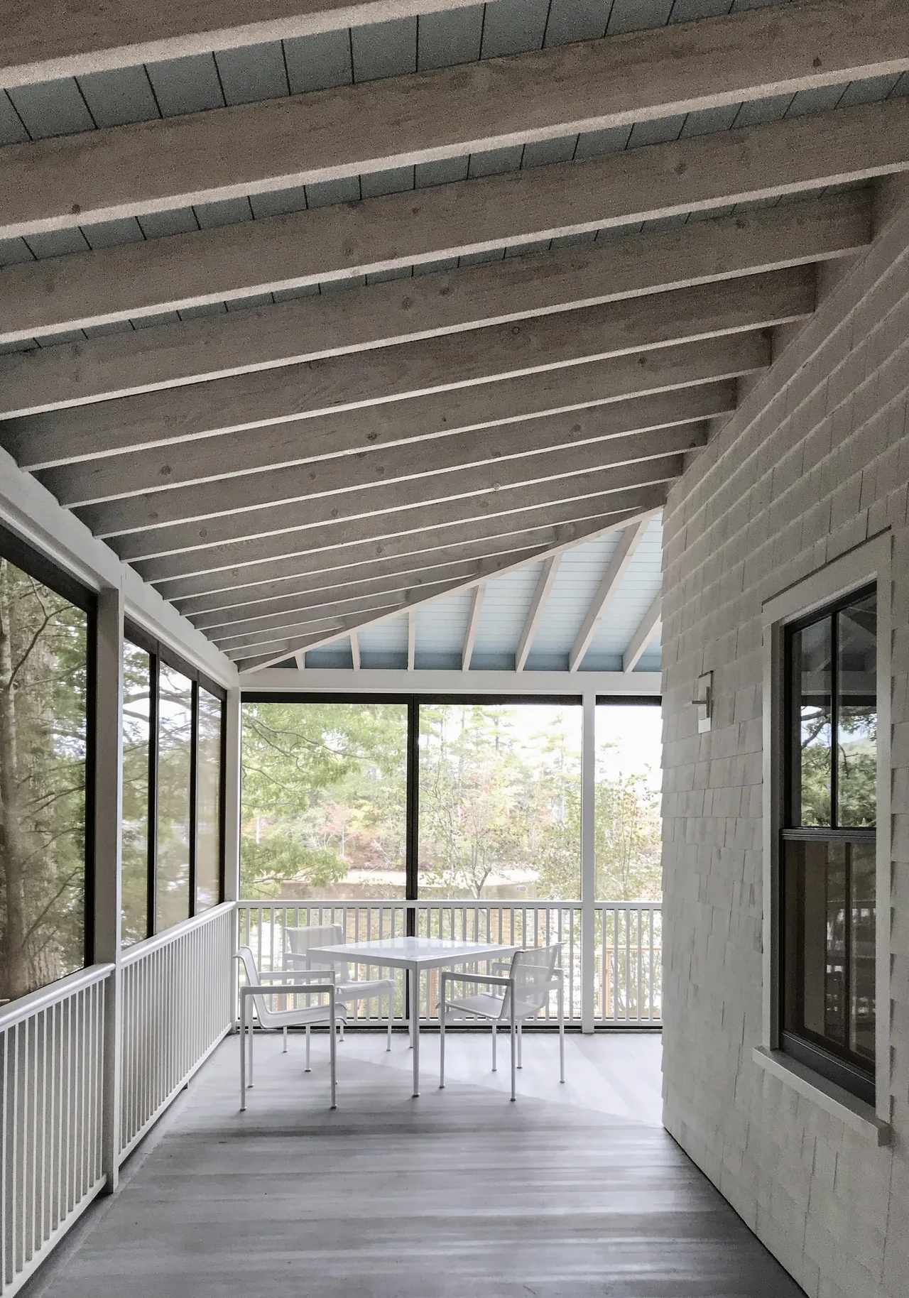 A screened porch at Camden Cottage features white wooden siding and simple outdoor furniture under an exposed beam ceiling.