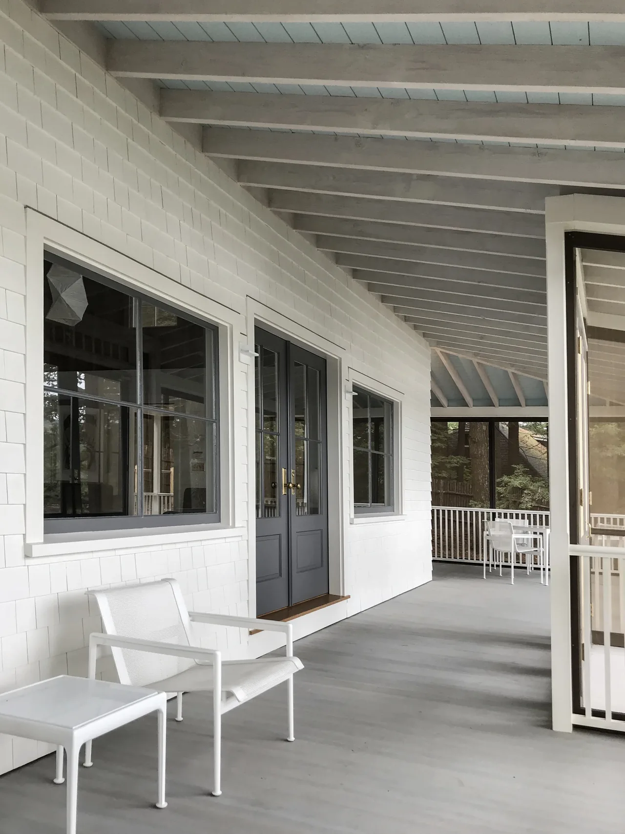The porch features a white shingle exterior, gray double doors with window panes, and white metal outdoor chairs and tables.