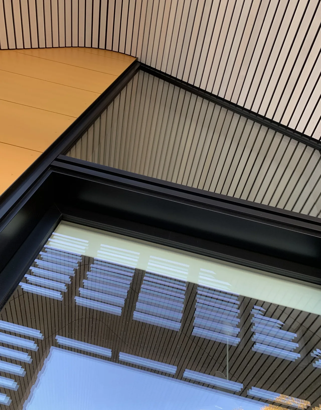Close-up of a modern building corner with vertical wood slats, black window frame, and yellow paneling reflecting on the glass.