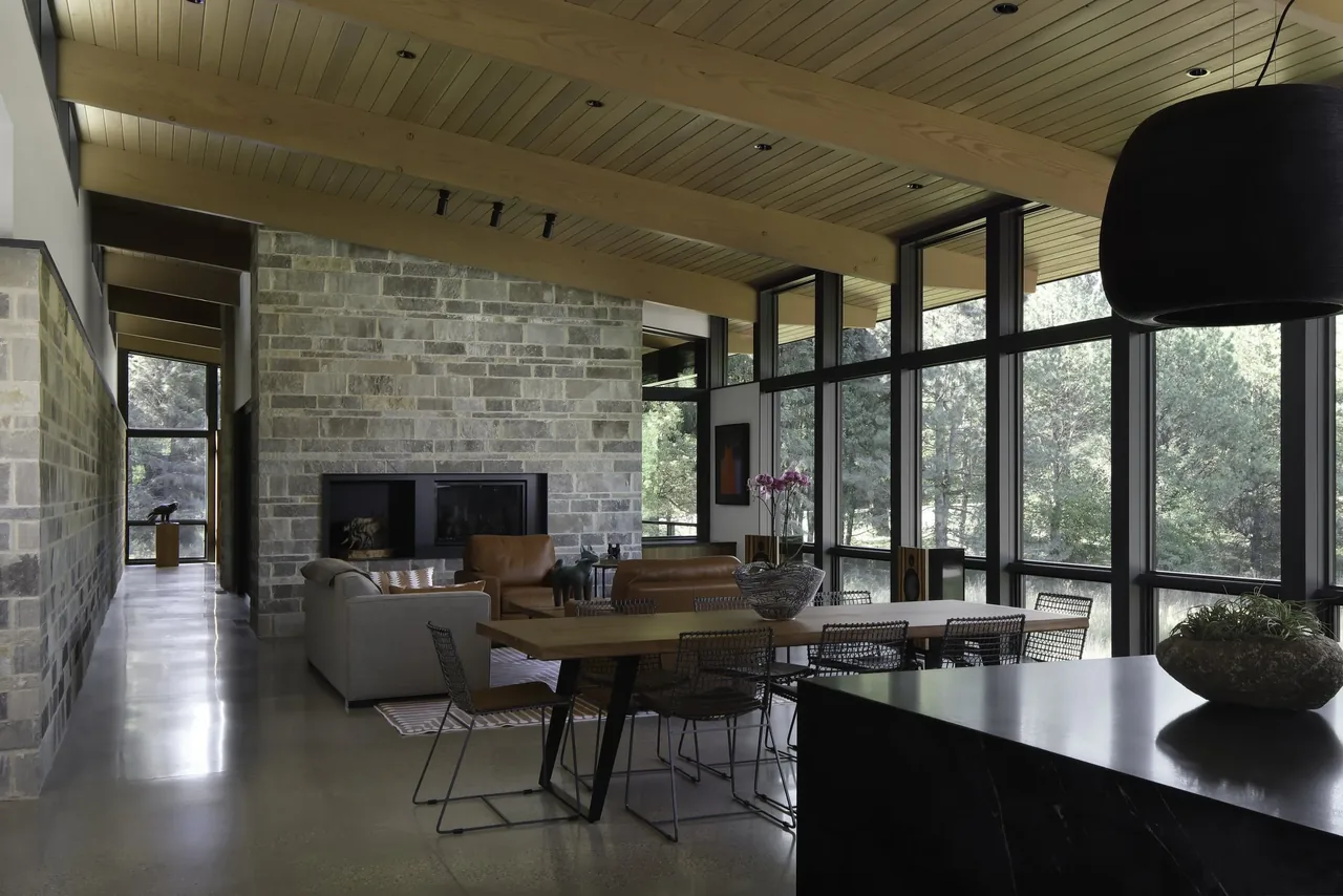 Living and dining area with floor-to-ceiling windows, stone fireplace wall, and exposed wood ceiling beams.
