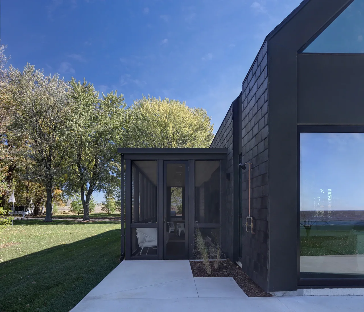 Exterior view of the Woven House featuring a black facade, large window, and a screened porch opening onto a lawn.