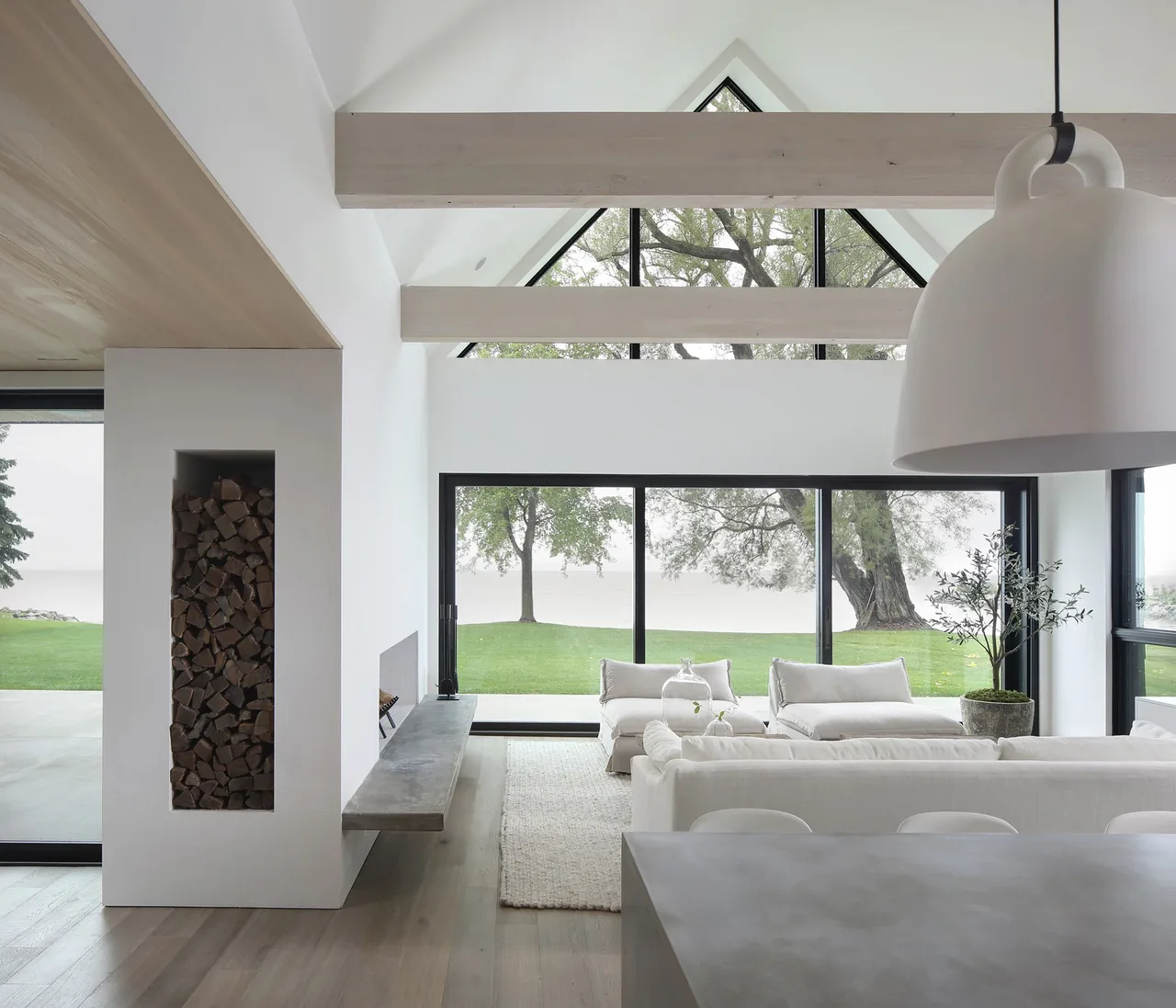 Interior of a living room featuring floor-to-ceiling windows with black frames, a floating concrete fireplace bench, and exposed ceiling beams.