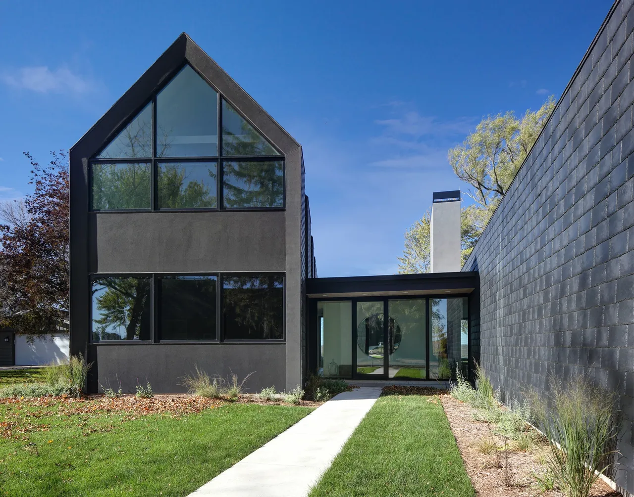 The Woven House features dark stucco and large black-framed windows with a gable roof and a glass entryway bridge.