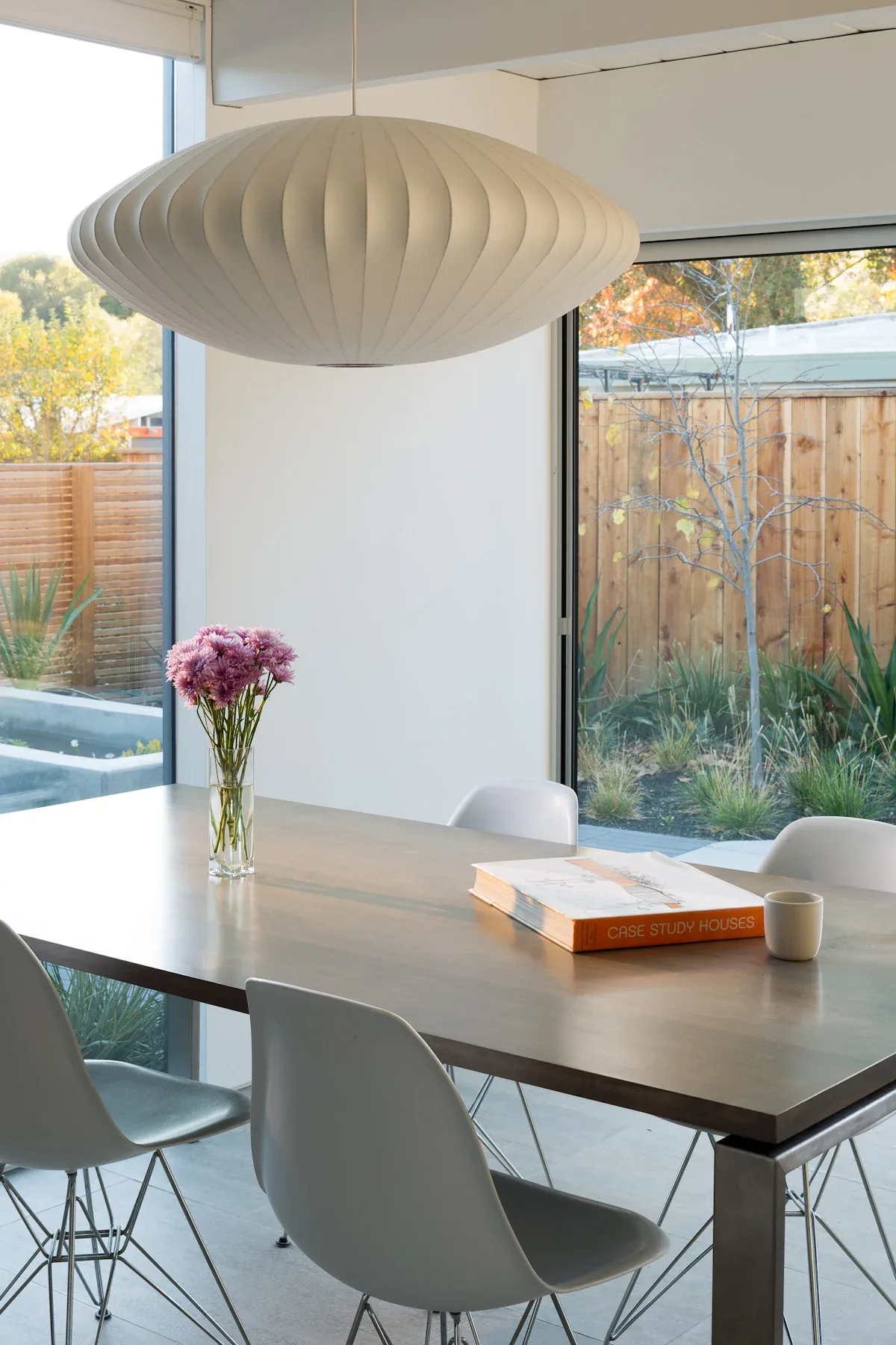 Dining space features a wood table and white molded chairs with large windows overlooking a garden and wooden fence.