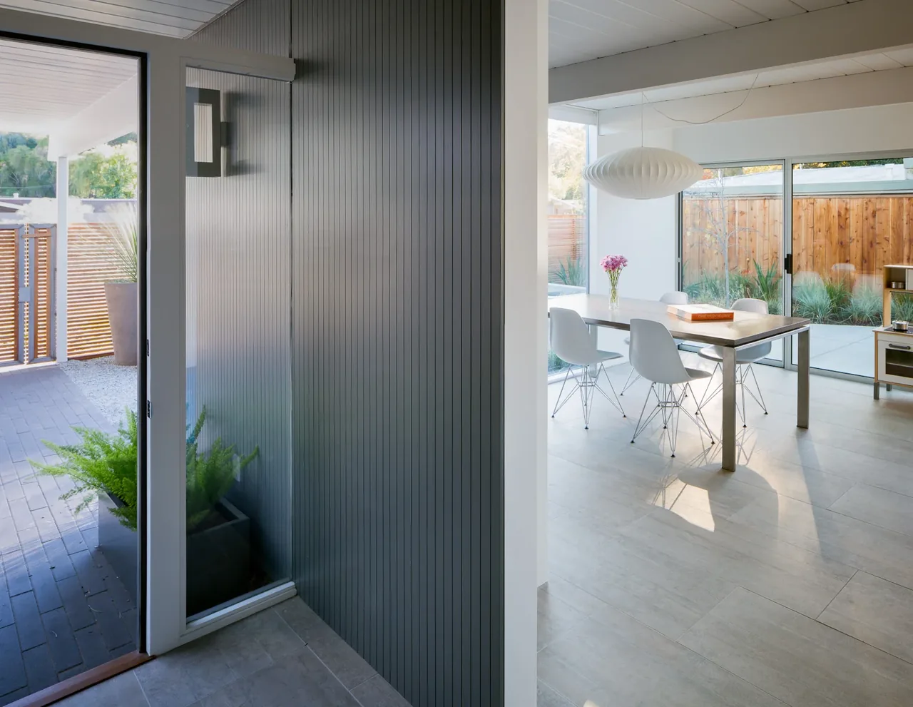 Dining room with white chairs and pendant light adjacent to an entry with vertical paneling and glass door.