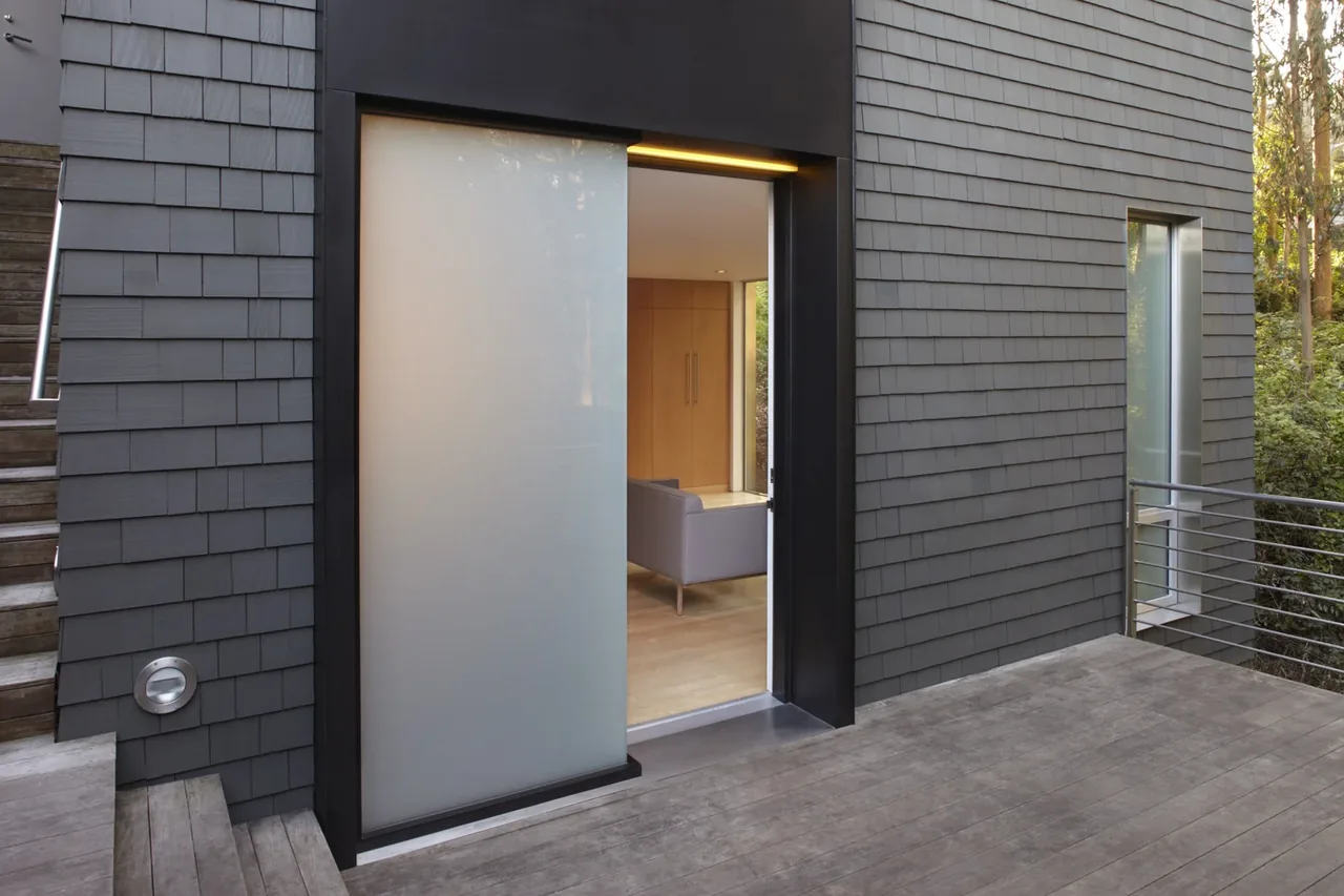 The Clarendon St. au pair suite features gray shingle siding with a sliding frosted glass door opening to a wooden deck.