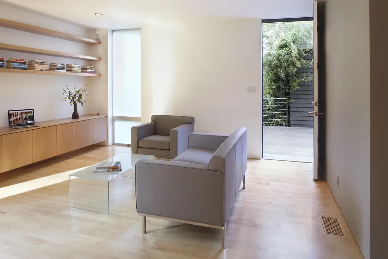 Minimalist living room in the Clarendon St. au pair suite shows floating wood cabinetry, pale wood flooring, and a glass coffee table.
