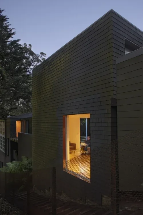 Modern dark-shingle clad building exterior with a large illuminated window showing interior space at dusk.