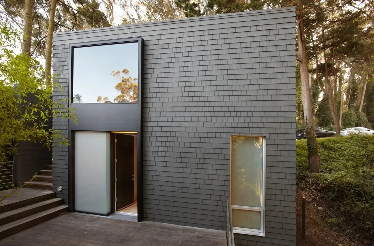 Exterior view of a modern gray shingle-sided two-story building with large windows and a black framed entrance door partially open.