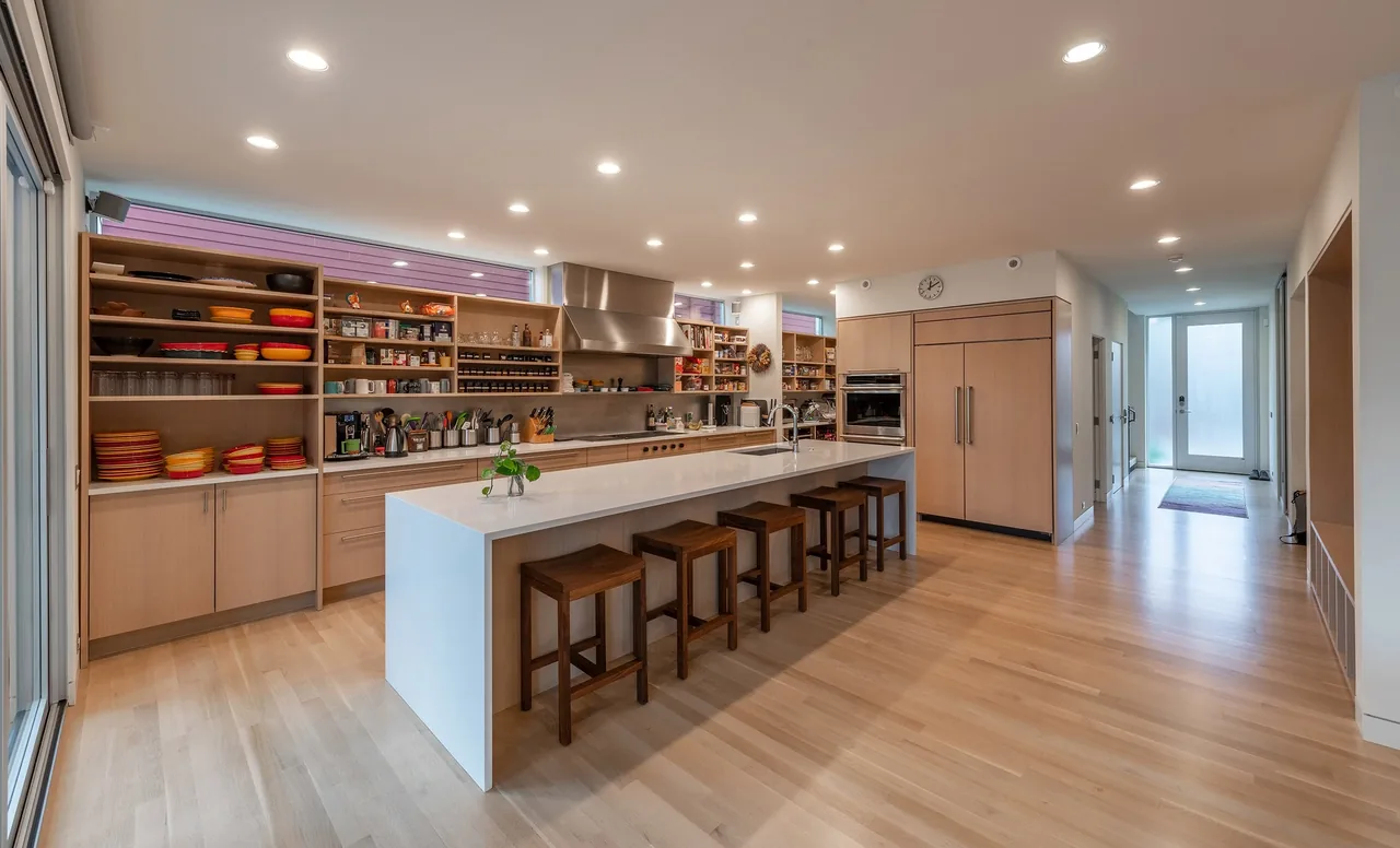 The Greenwood Residence kitchen features a minimalist design with light wood cabinetry, a large island, and open shelving for dish storage.