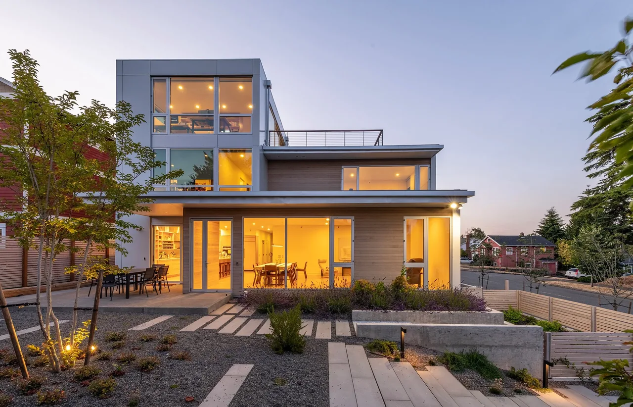 The Greenwood Residence features a modern exterior with large glass windows and landscaped front yard lighting in the evening.
