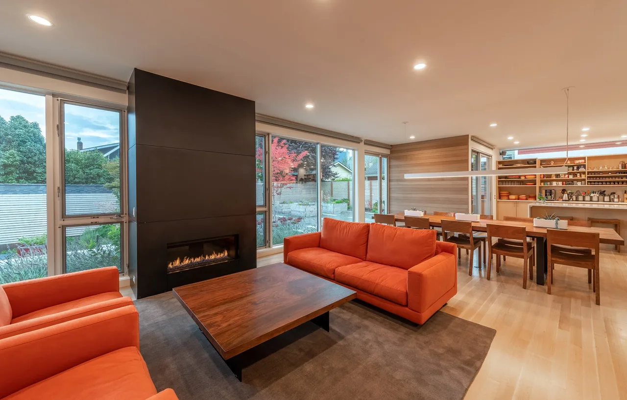 The Greenwood Residence living and dining area features orange couches, a modern fireplace, and large windows overlooking the landscaped yard.