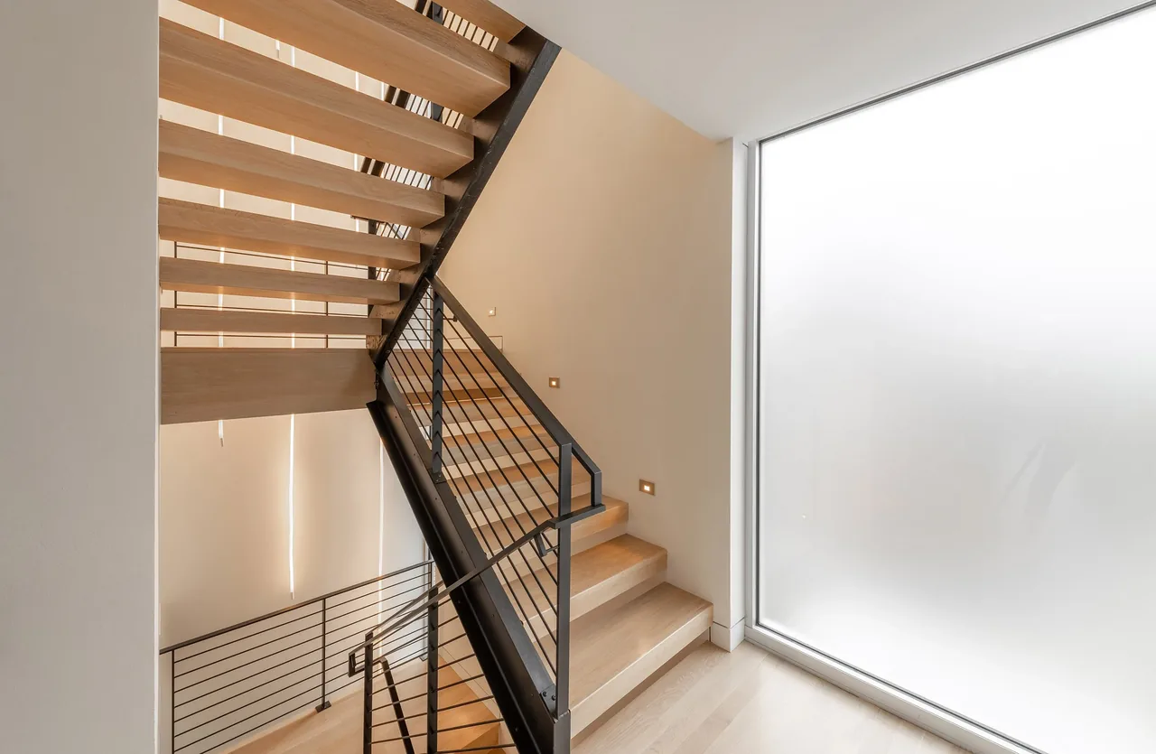 Modern stair with black metal railings and wood steps at Greenwood Residence, featuring a large frosted window.