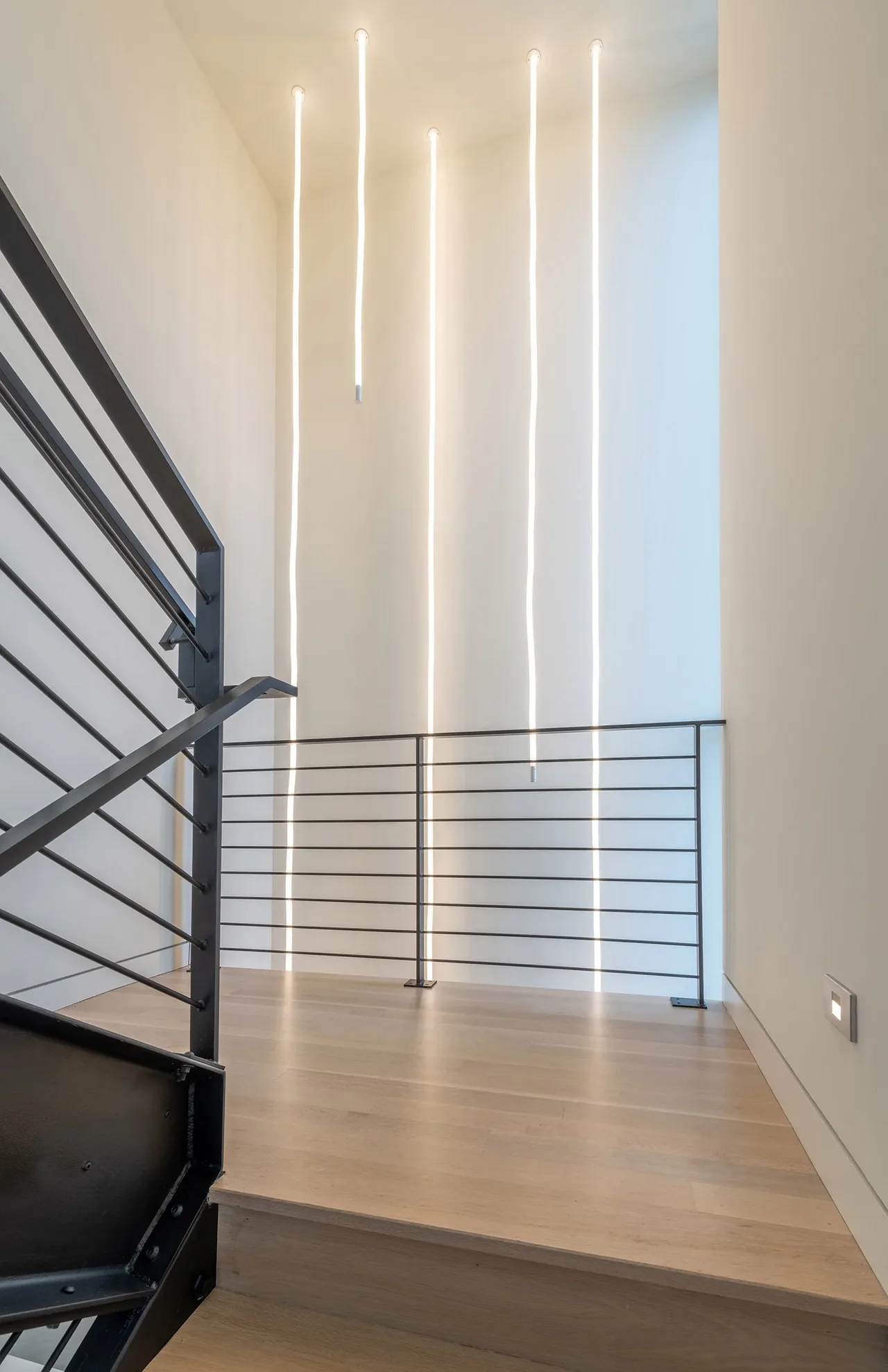 A stair landing features horizontal metal railings and four slender vertical pendant lights.