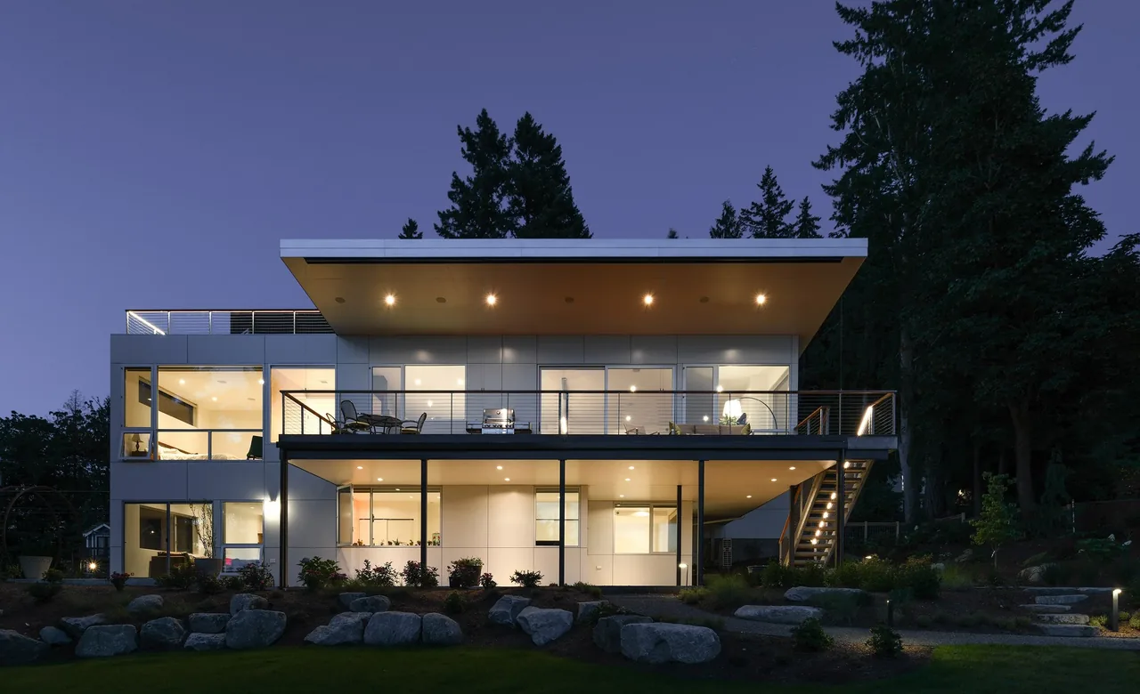 Sammamish Residence features a modern flat roof and expansive glazing with an elevated outdoor deck and stairs, set against a wooded backdrop.