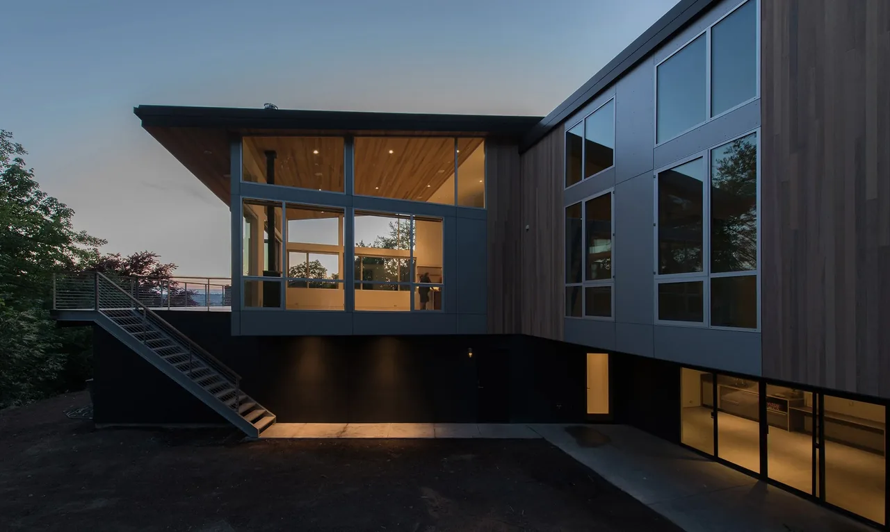 Mercer Island Residence by Build LLC features large aluminum windows and wood siding at dusk.