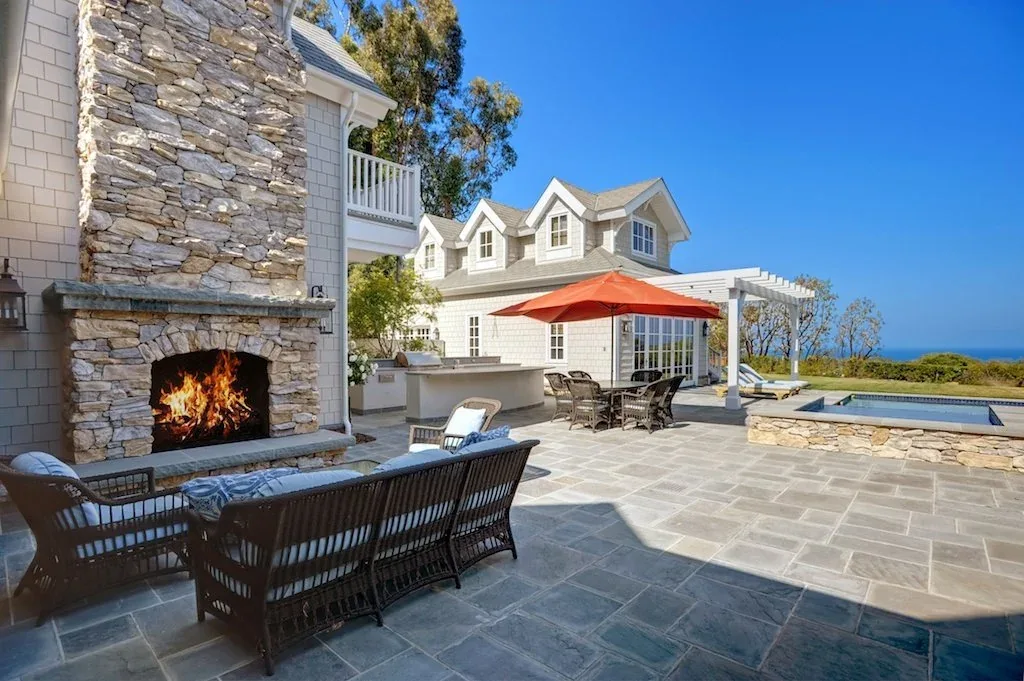 Outdoor patio featuring a stone fireplace, wicker seating, dining area with umbrella, and a pool with ocean views at the Dume Drive Traditional project.