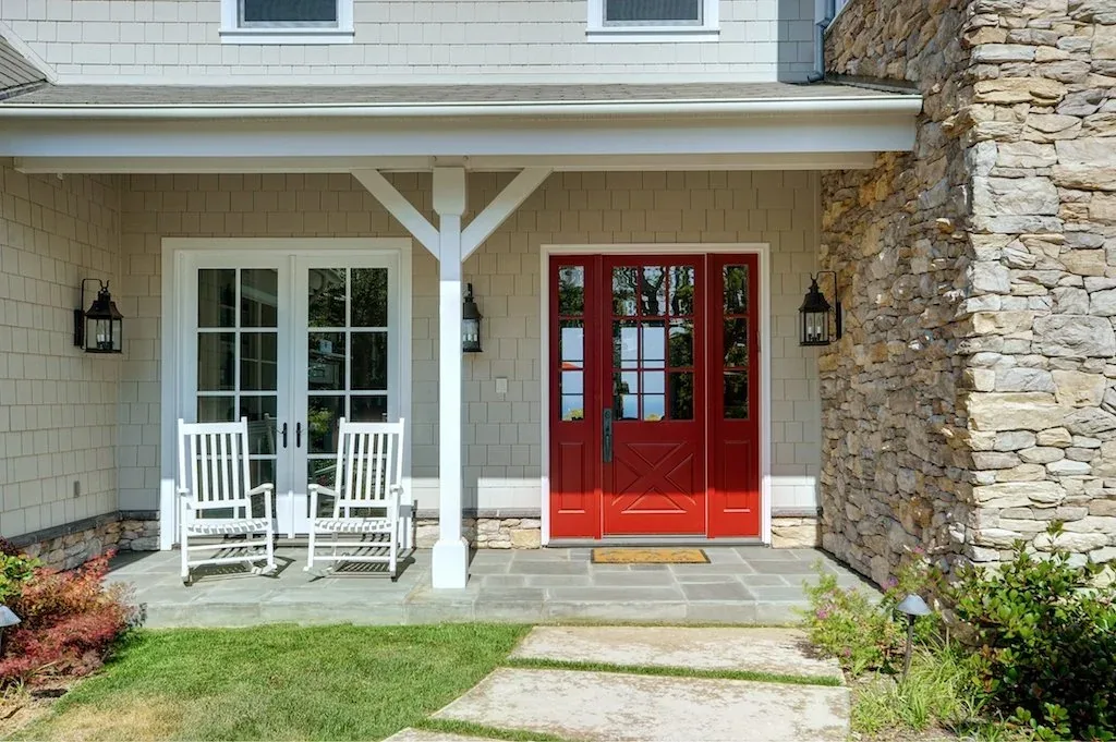 Traditional front porch with a bold red entry door, stone wall detail, and white rocking chairs under a covered patio.
