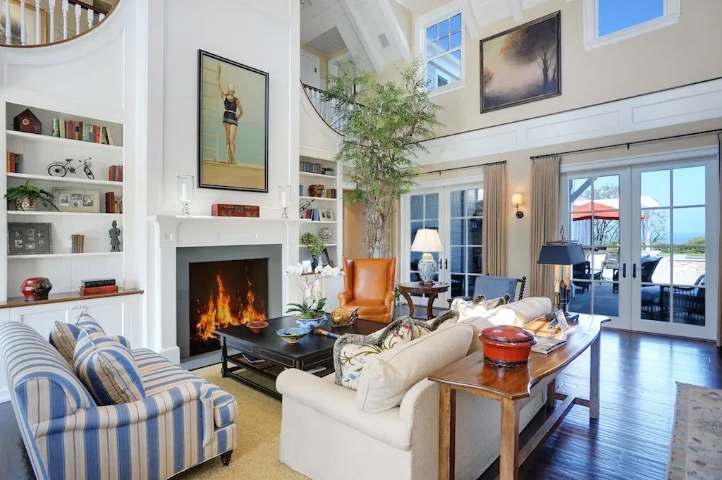 Dume Drive Traditional living room with double-height ceiling, fireplace, and French doors opening to a patio with umbrellas.
