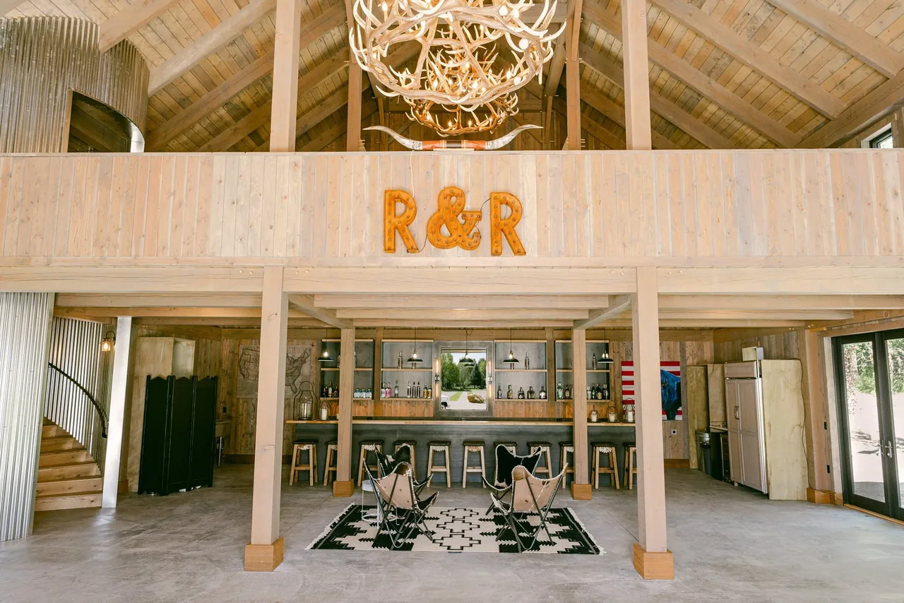 Open living space with timber framing, a large antler chandelier, and a bar area under a loft with wood paneling at Riverside Retreat.