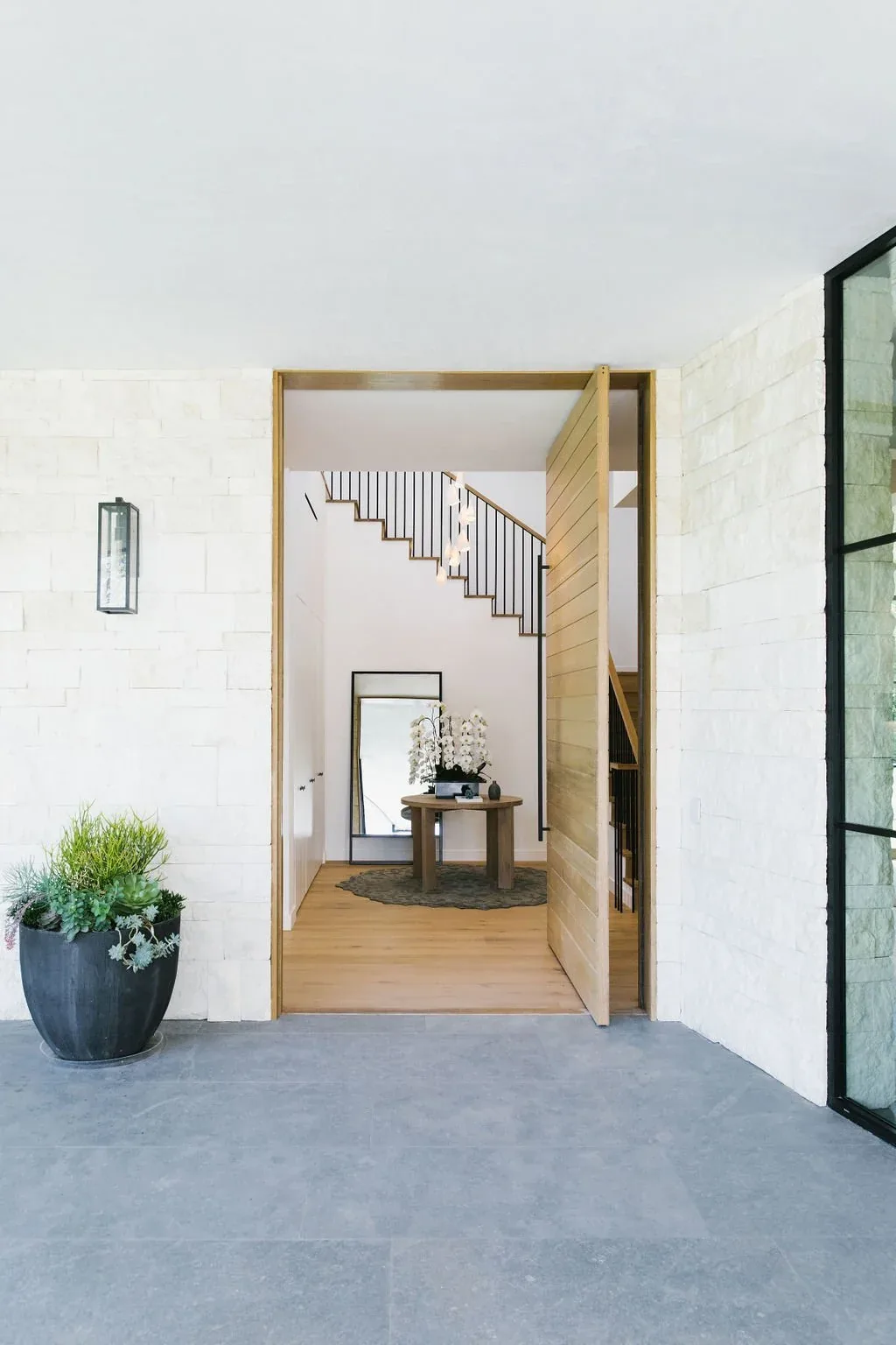 The Malibu Modern foyer features a large pivot wooden door opening to a minimalist interior with stairs and decorative table.