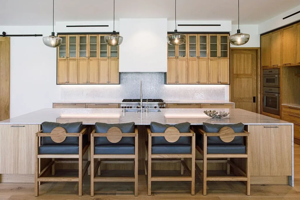 The Malibu Modern kitchen features warm wood cabinets, a large marble island, pendant lighting, and built-in ovens.