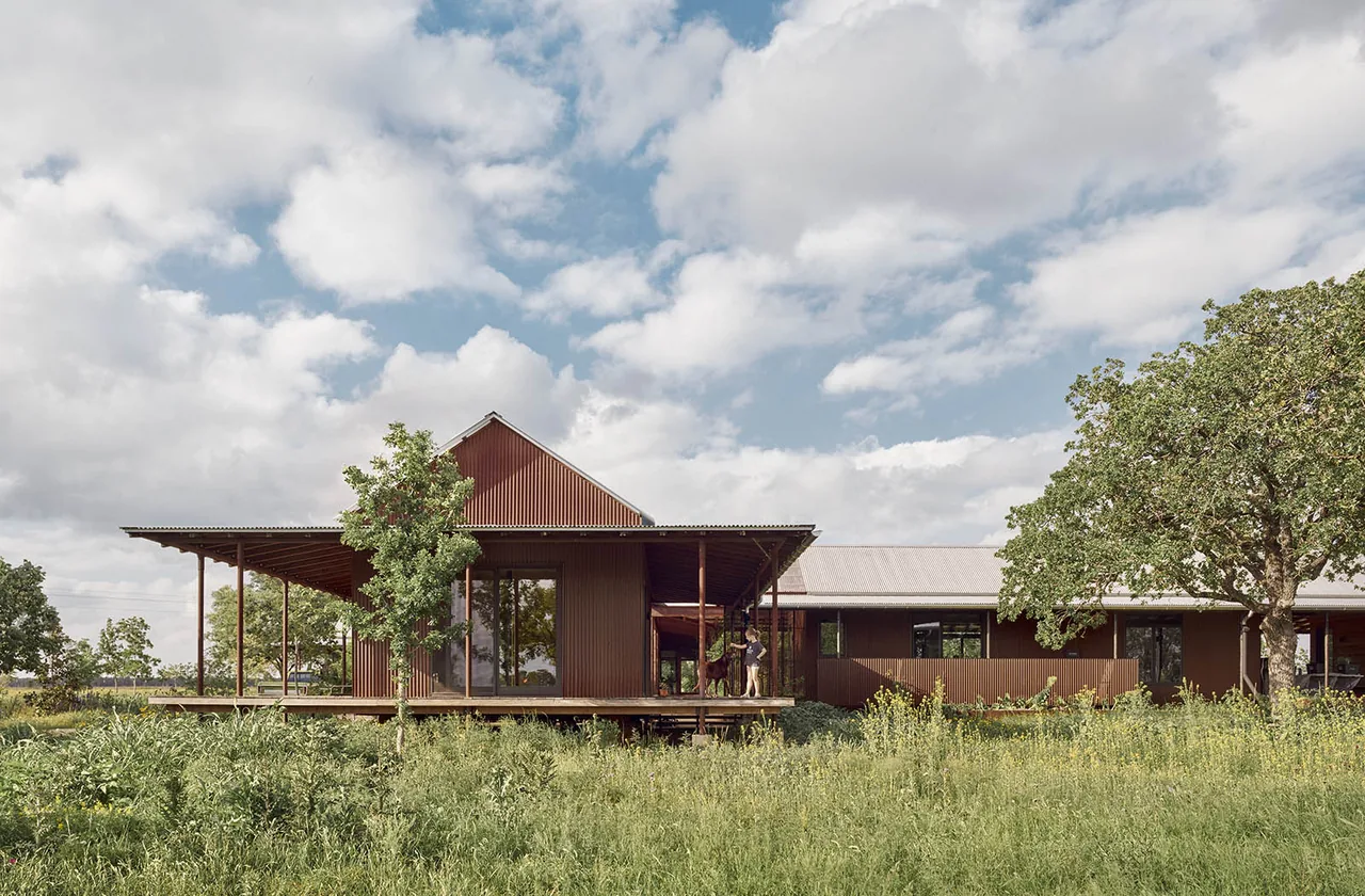 Roam Ranch by Burton Baldridge Architects features rust-colored siding with a raised foundation and covered porch in a rural setting.