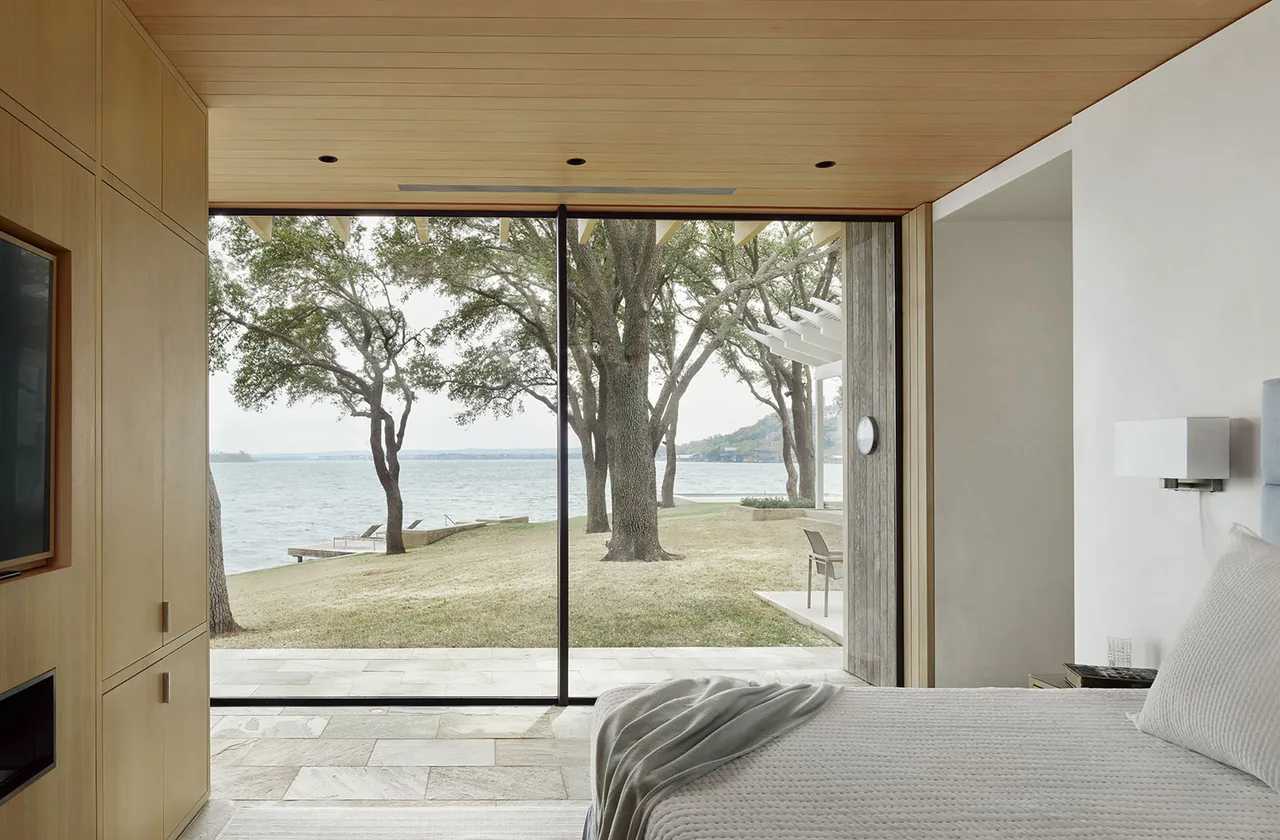 Lakefront bedroom with floor-to-ceiling sliding glass doors opening to a stone patio and lakeside view.