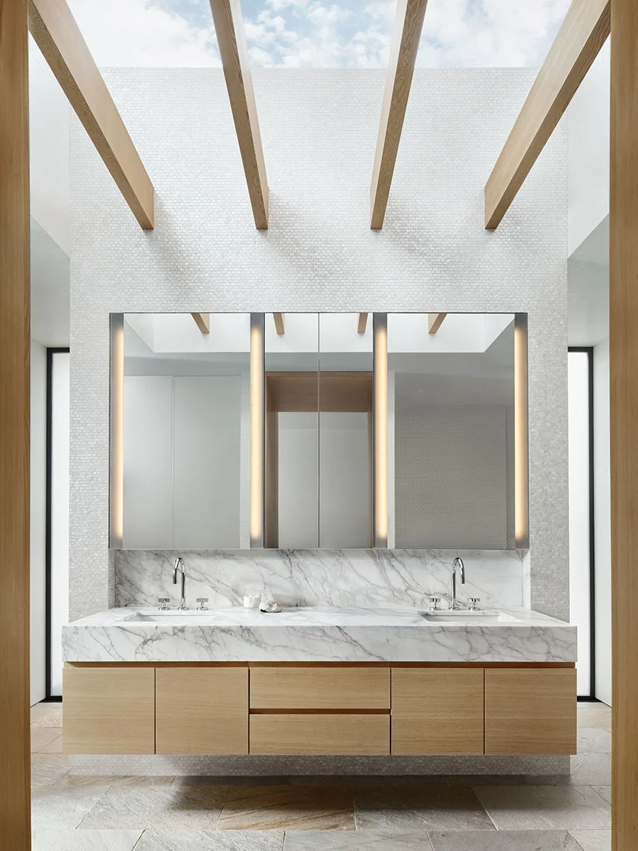 Modern bathroom vanity features a marble countertop, double undermount sinks, and vertical lighted mirrors beneath exposed wooden beams.