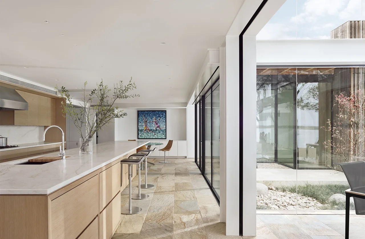 Modern kitchen featuring a marble island with stools and floor-to-ceiling sliding glass doors opening to a courtyard with stones and plants.