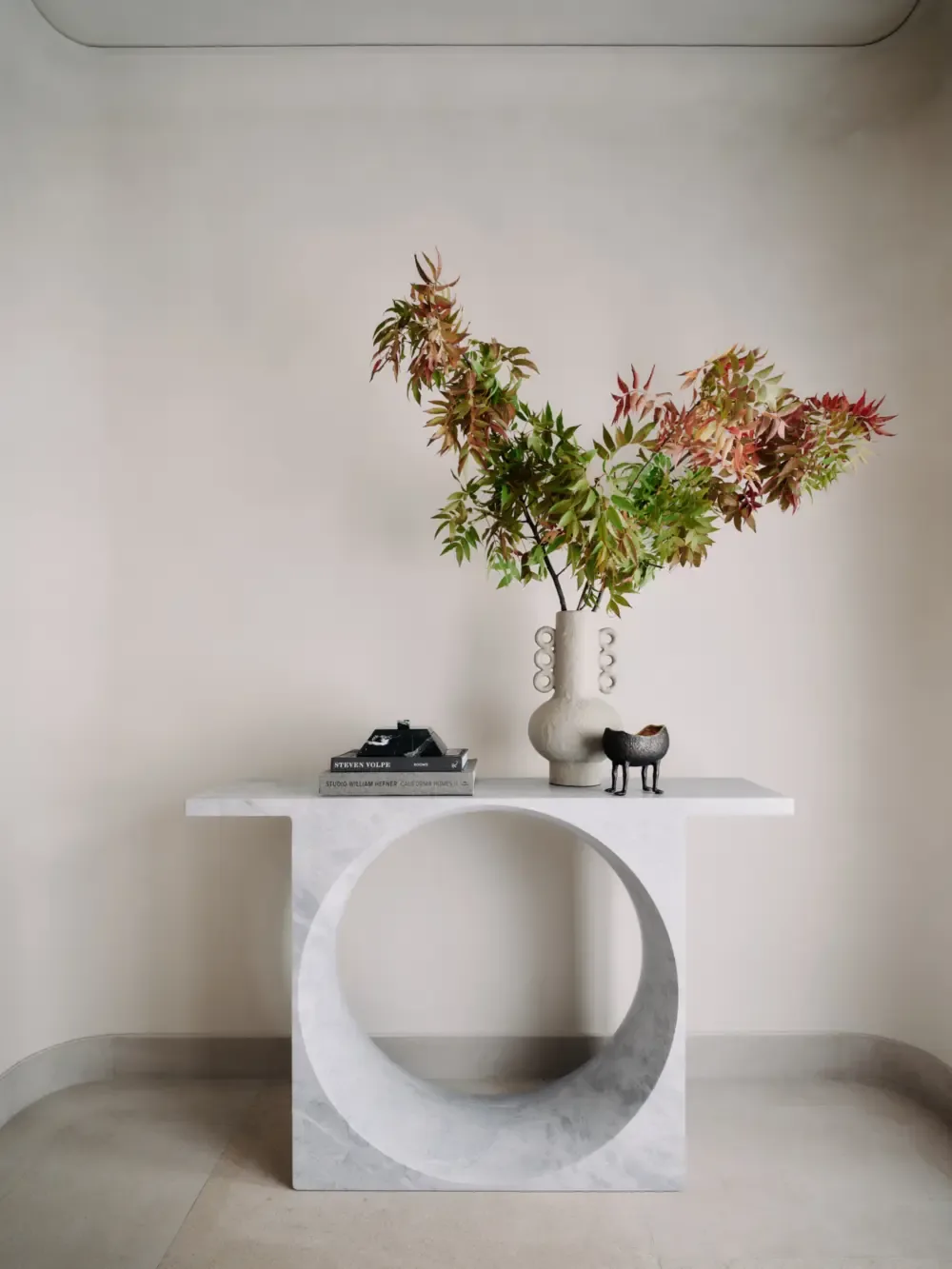 A white marble console table with a circular cutout is styled with a sculptural vase, books, and a small animal figurine.