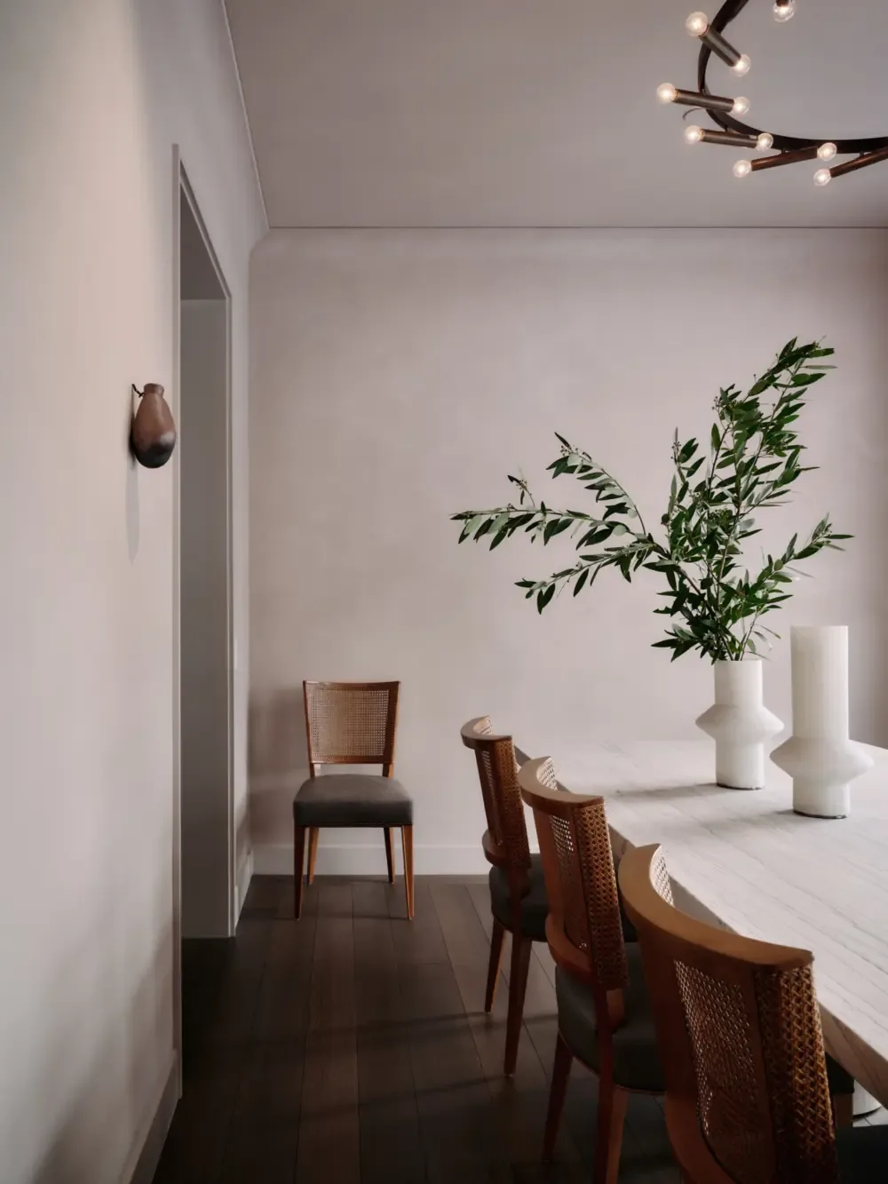Dining room featuring wooden chairs with woven backs, a white marble table, and minimalist ceramic vases with greenery.