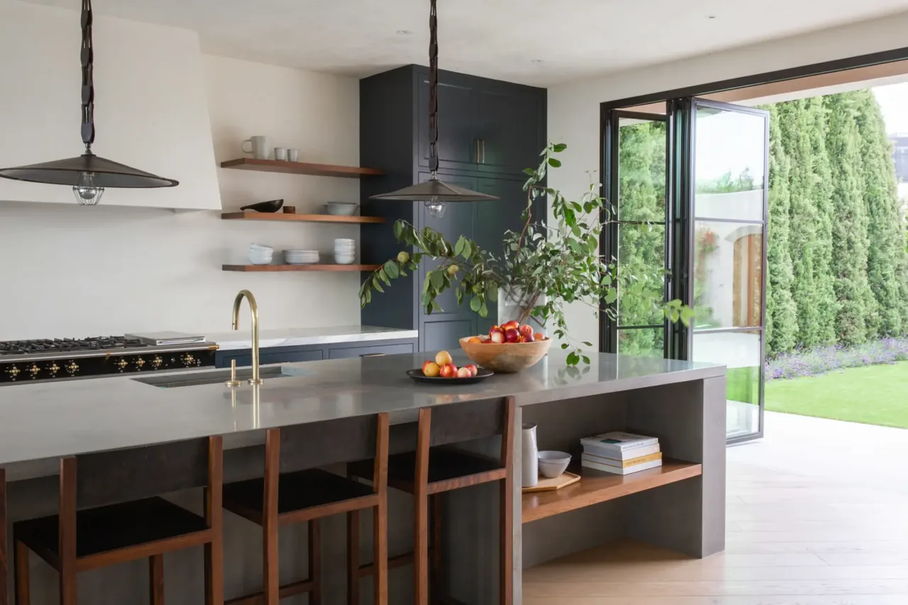 Modern kitchen features a gray island with wooden bar stools, brass fixtures, open wooden shelves, and black metal framed folding doors.