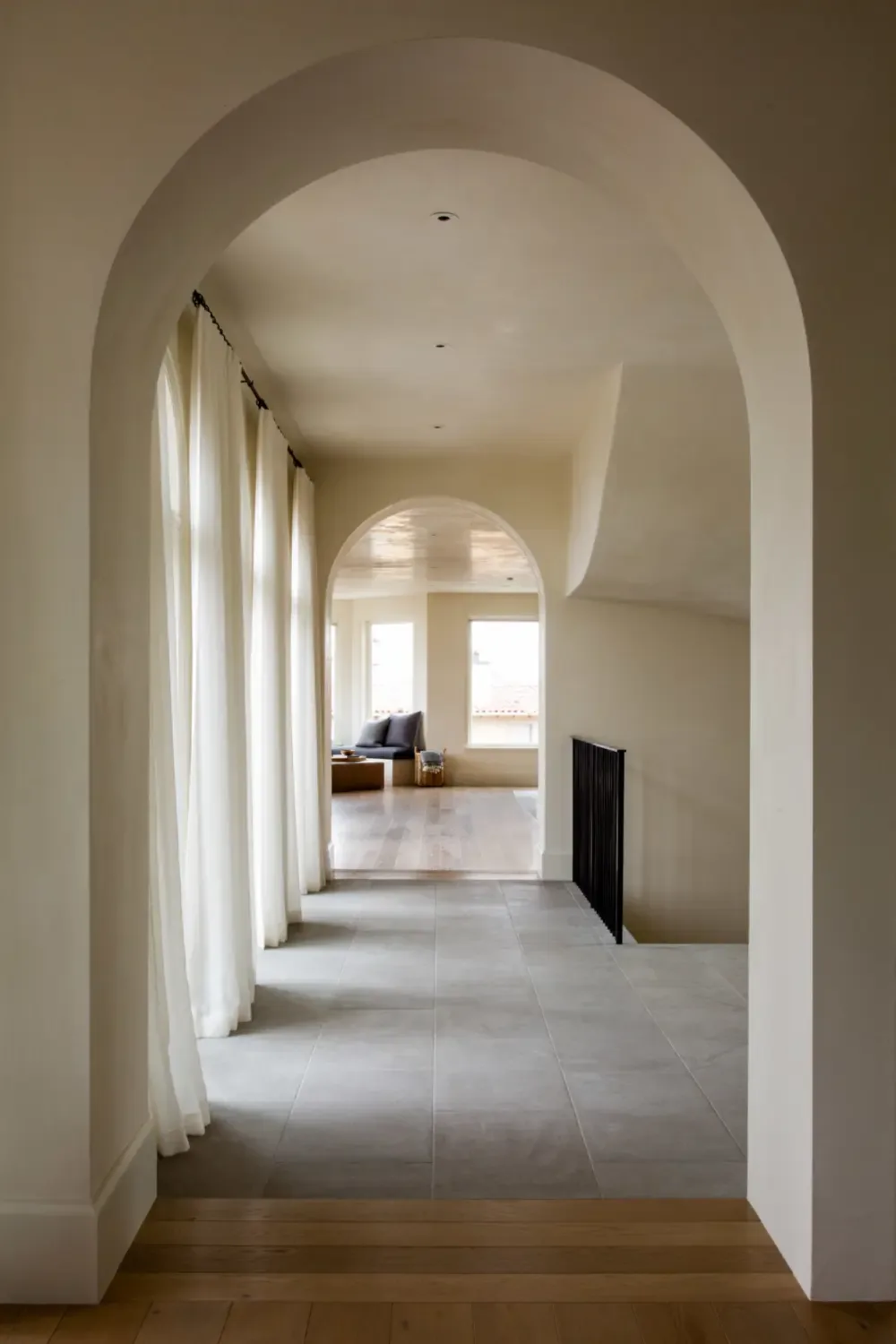 Hallway with arched openings and sheer curtains leading to a sunlit living space with neutral tones.