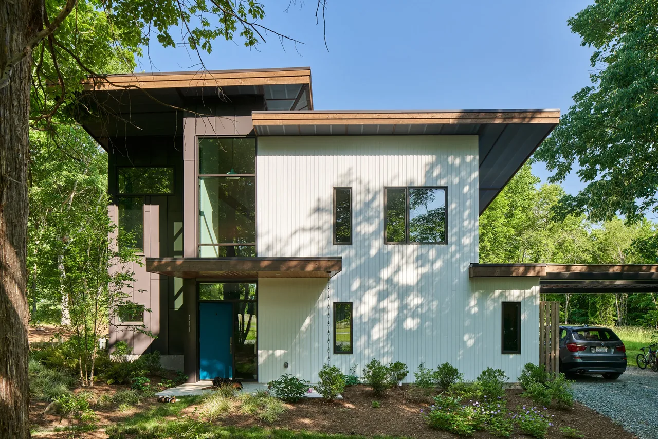 Acorn Acres features a modern two-story home with white vertical siding, flat and shed roofs, and large windows in a wooded setting.