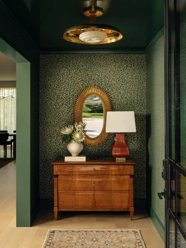 Entry hallway features vintage wood dresser topped with a lamp, decorative vase, and sunburst mirror on floral wallpaper.