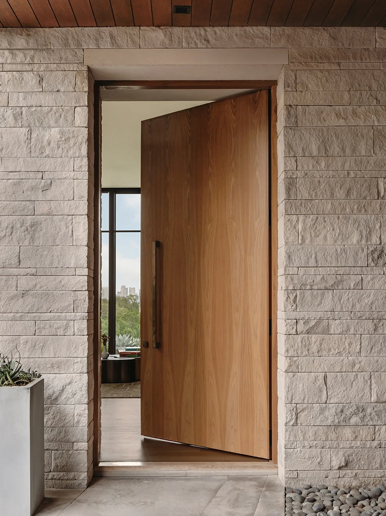 Rollingwood Modern's front entrance features a pivot door framed by textured stone walls and concrete flooring.