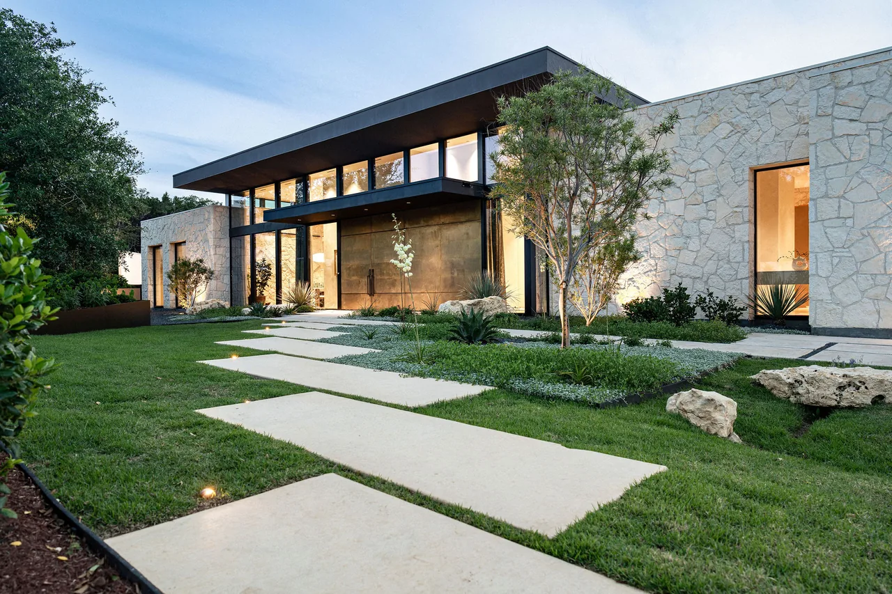 Modern residence features stone walls, flat roof, large black-framed glazing, and landscaped front yard with stepping stones in Austin.