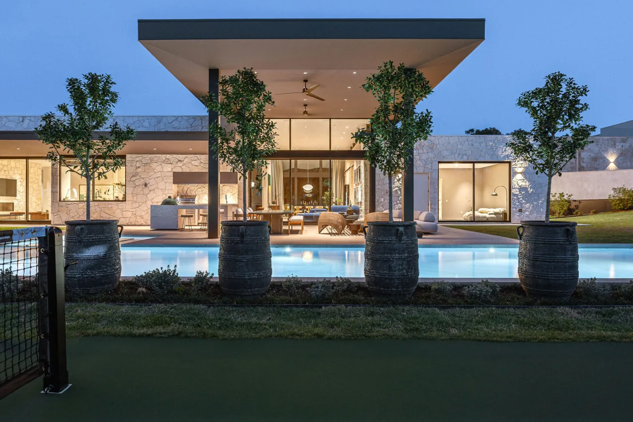 Modern stone-sided house with covered patio, outdoor dining, and pool illuminated at dusk.