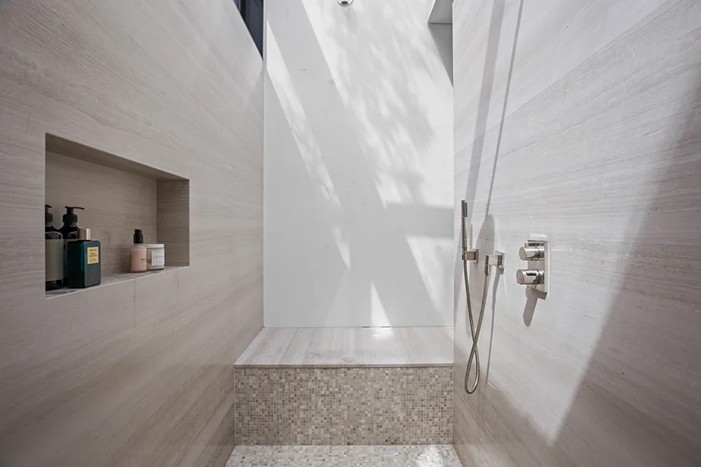 Bright shower area features a built-in niche, mosaic tiled bench, and modern chrome fixtures with natural light casting shadows.
