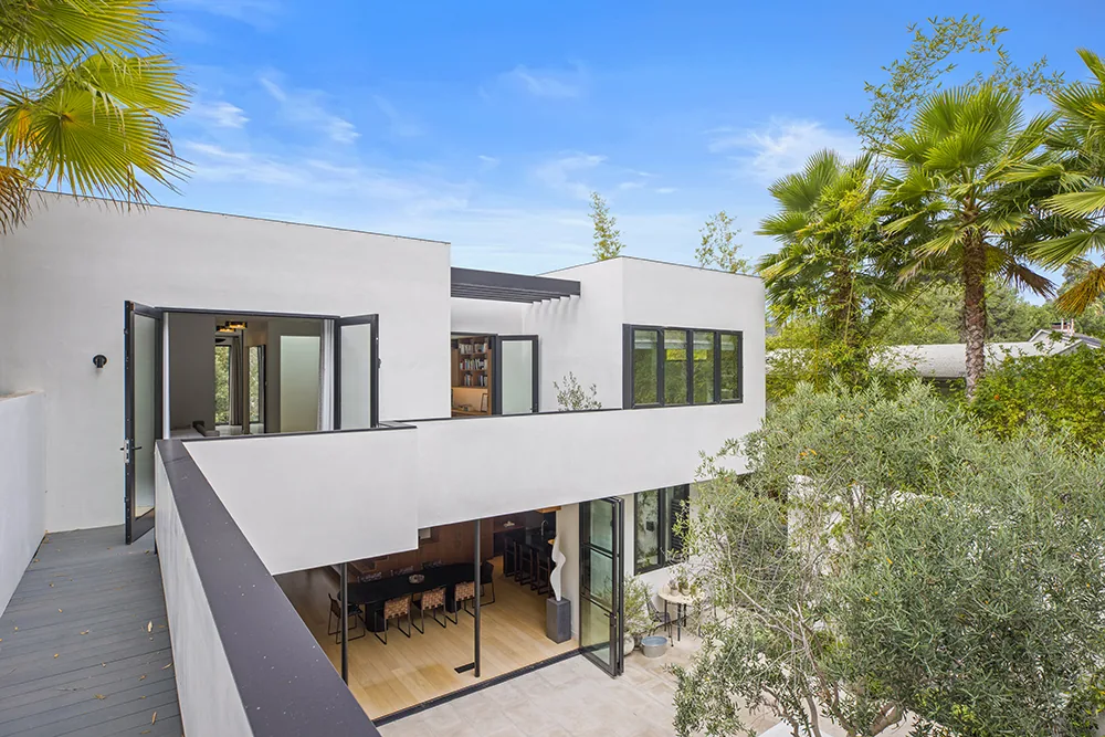 Halo House features a modern white stucco exterior with balcony and floor-to-ceiling glass doors opening onto a patio with greenery.