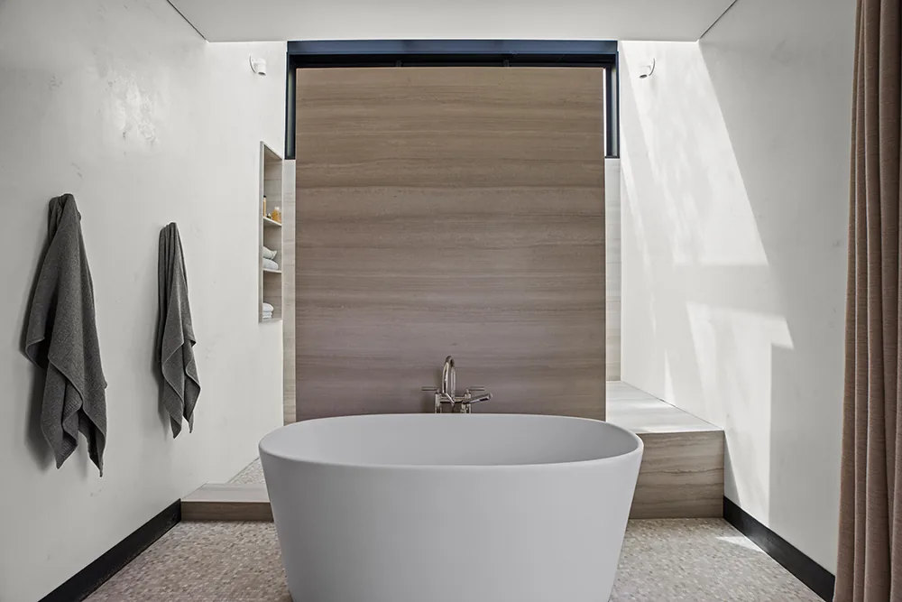 The Halo House bathroom features a freestanding tub with a wood-paneled wall and natural light from a clerestory window above.