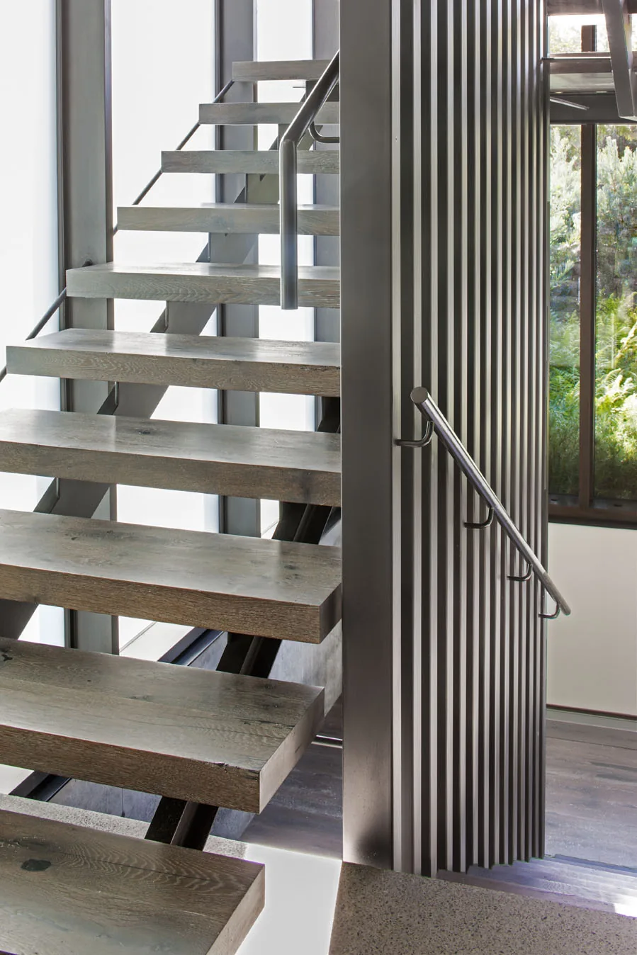 Floating wood stairs with metal handrails and a black vertical slat partition near a window.
