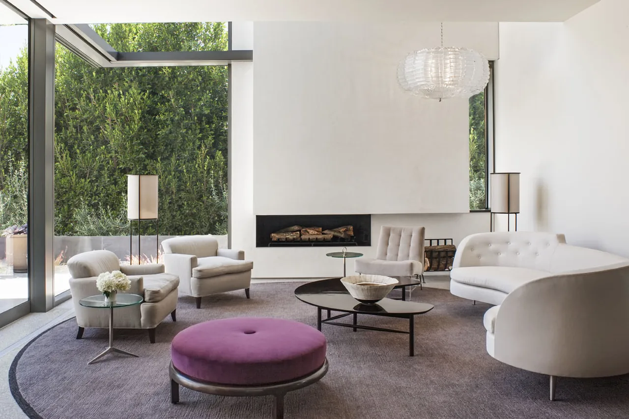Living room features a fireplace, white walls, large windows, and a glass side table paired with contemporary white armchairs and sofa.