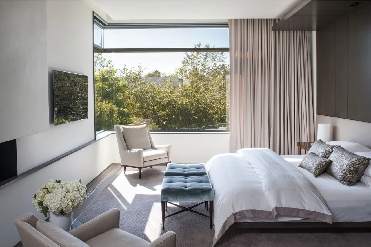 A bright bedroom with a large corner window providing views of lush trees outside.
